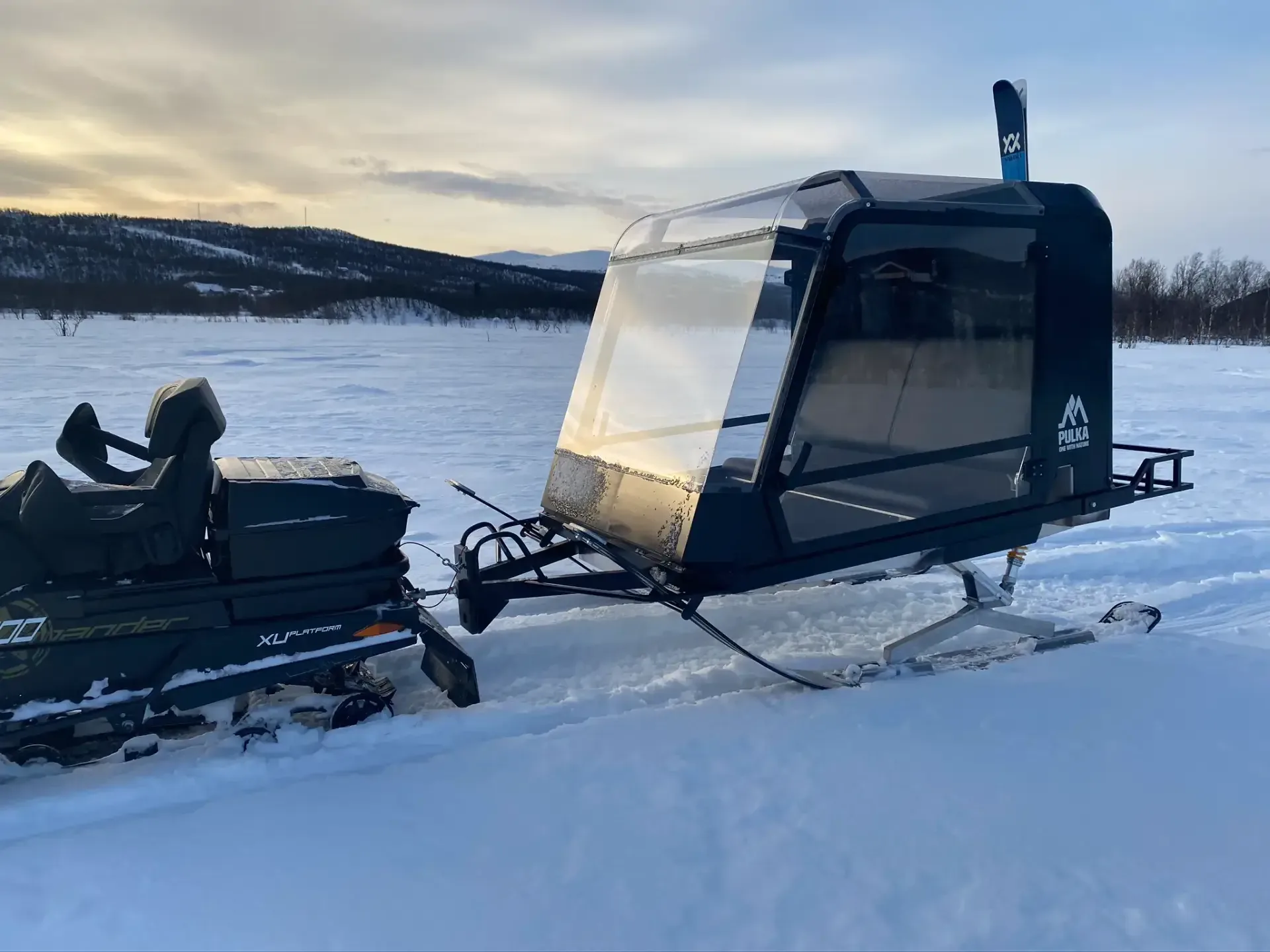 Pulkapanorama parkerad på en frusen sjö i solnedgång, med fjäll i bakgrunden