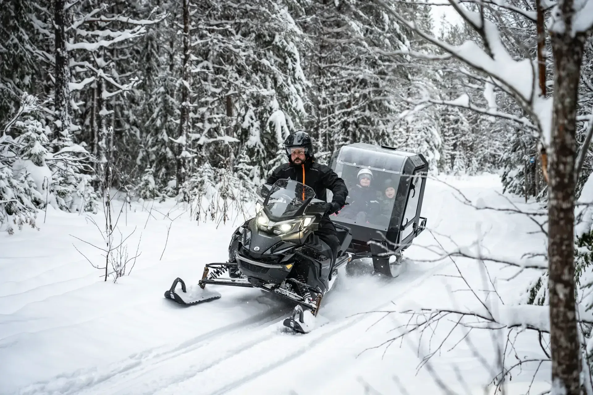Pappa kör skoter med Pulkapanorama genom snötäckt skog, två barn syns genom panoramafönstret